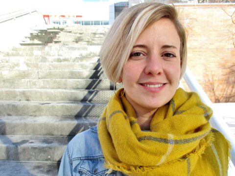 Smiling woman with short, blonde hair and a yellow scarf stands near steps in the cold.